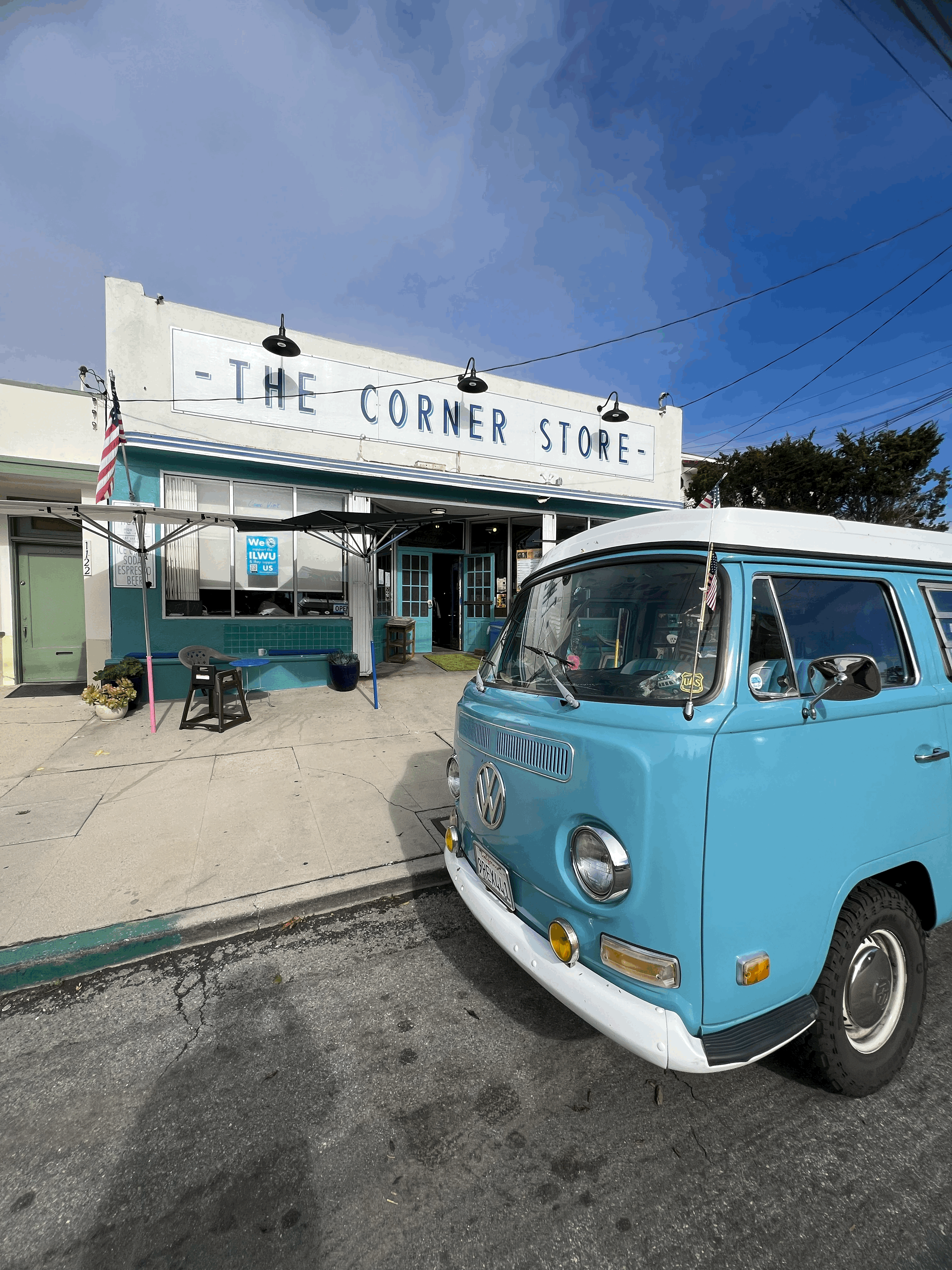 Exterior of The Corner Store café with a vintage blue Volkswagen van parked out front, showcasing a retro neighborhood restaurant vibe under a bright blue sky in coastal California.