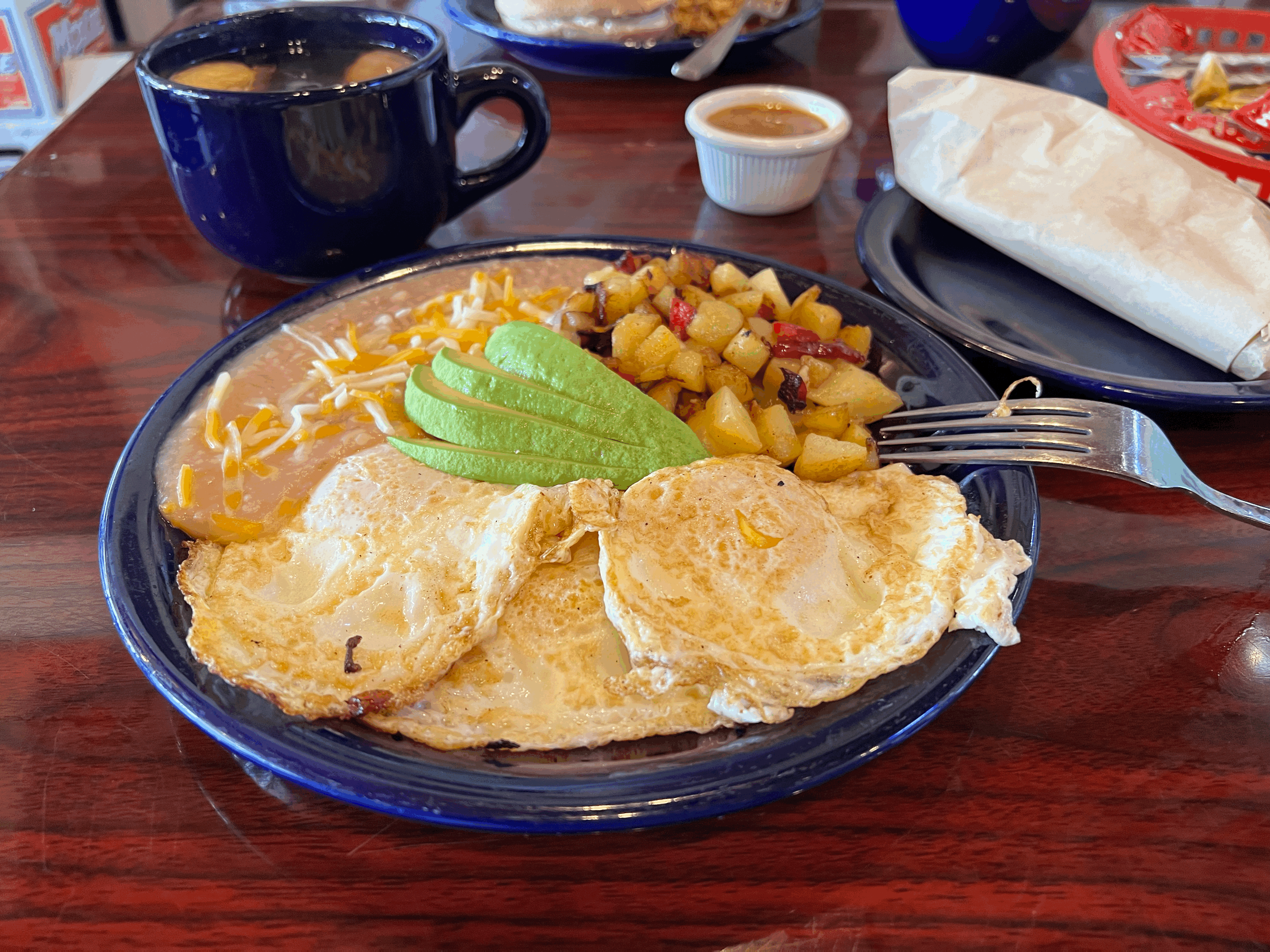 Classic breakfast plate with sunny-side-up eggs, sliced avocado, seasoned breakfast potatoes, and refried beans topped with melted cheese, served with a cup of coffee at a cozy diner-style café.