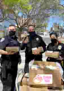 3 Santa Monica police, two men and one woman, standing outside with black masks each holding a box lunch in front of a cart full of Urth Caffé lunch boxes. Thank You is at bottom right.