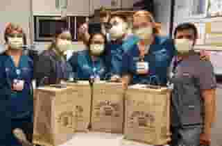 Group of six nurses and doctors wearing masks and blue scrubs standing behind 4 large Urth food bags on table. Male nurse in background is giving thumbs up.