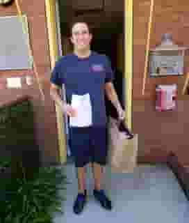 Santa Monica Fireman in blue T-shirt and shorts holding large Urth Caffé bag of lunch boxes. He is standing just outside a narrow doorway leading into the firestation.