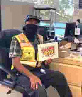 Man sitting at desk with black mask, hat, yellow vest and holding a thank you sign.