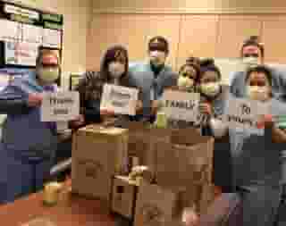 7 White Memorial Hospital nurses with yellow masks holding four sheets of paper that say "Thank You From our Family To Yours" with image of red heart. They are standing behind a table full of Urth Caffé food bags and coffee.