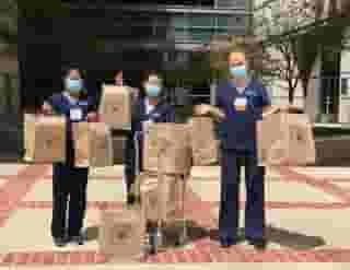 Three nurses with blue masks standing in front of the Ronald Reagan UCLA Med center holding multiple large Urth food bags and a cart with bags in hospital courtyard.