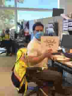 Man sitting at desk with mask and holding a thank you sign. In the background is another man is sitting at another desk with mask and holding the Urth Caffe "To Our Brave Heroes" card.