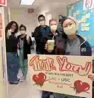 Five nurses standing in a hospital hallway wearing yellow medical masks and holding a green Urth Caffé coffee cups. Front nurse is holding a sign with two big red hearts that reads: Thank You! From the HEART LAC + USC Emergency Room Staff.