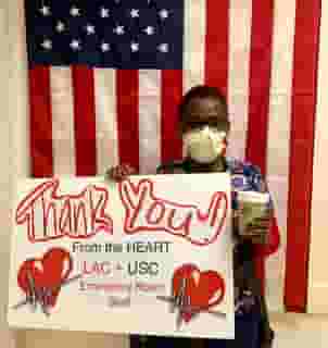 One USC nurse wearing yellow medical mask holding a sign with two big red hearts that reads: Thank You! From the HEART LAC + USC Emergency Room Staff. She is also holding a green Urth Caffé coffee cup and standing in front of a vertical USA flag.