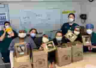 Group of nurses with masks in room with 4 Urth bags and 4 lunch boxes on table with dry erase white board behind them that say, Thank You Urth Caffé!