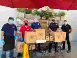 Two Urth employees and three Beverly Hills firemen standing behind table with Urth bags of lunch boxes and coffee. Rock wall fountain with flowers in background.