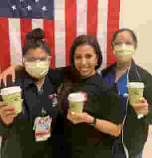 3 nurses holding green Urth Caffé To Go coffee cups. They are standing in front of a vertical USA flag, two with masks and protective eye wear and the one without a mask is smiling.