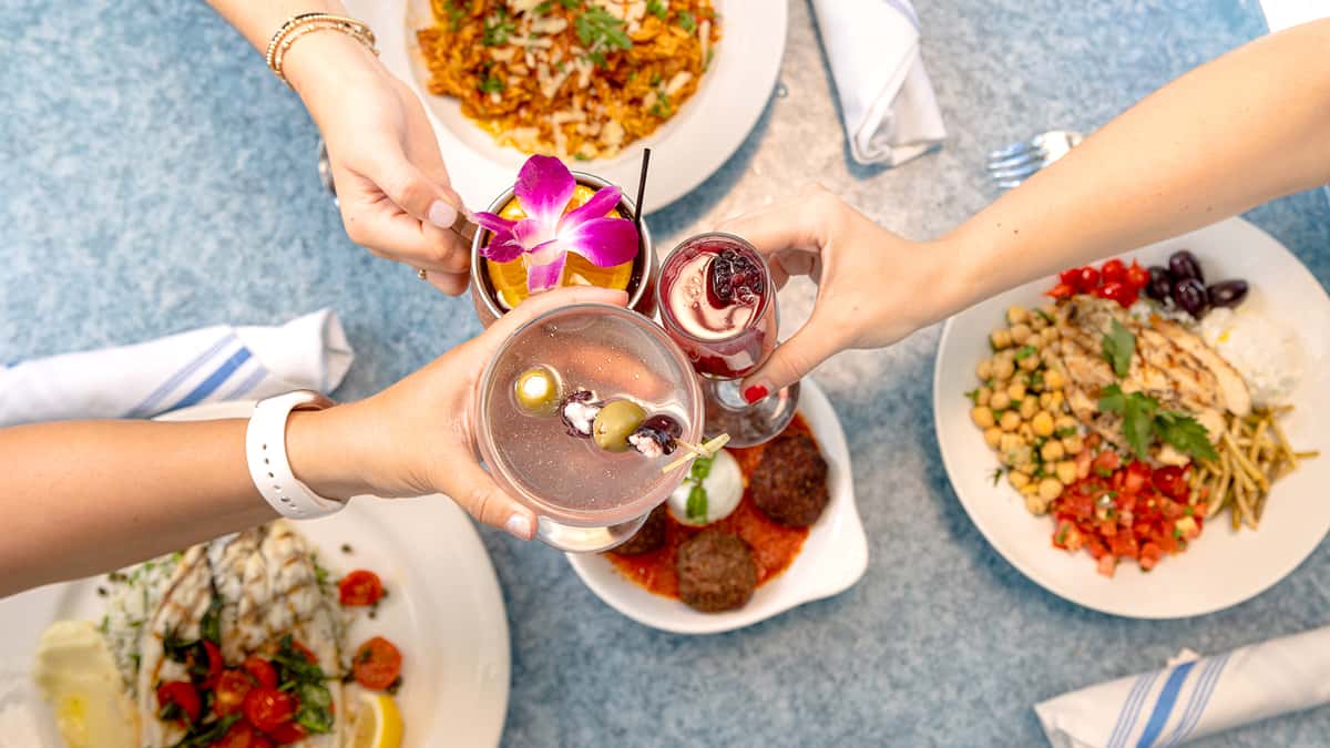 Guests cheersing glasses over a group shot of food on a table at Taverna Agora