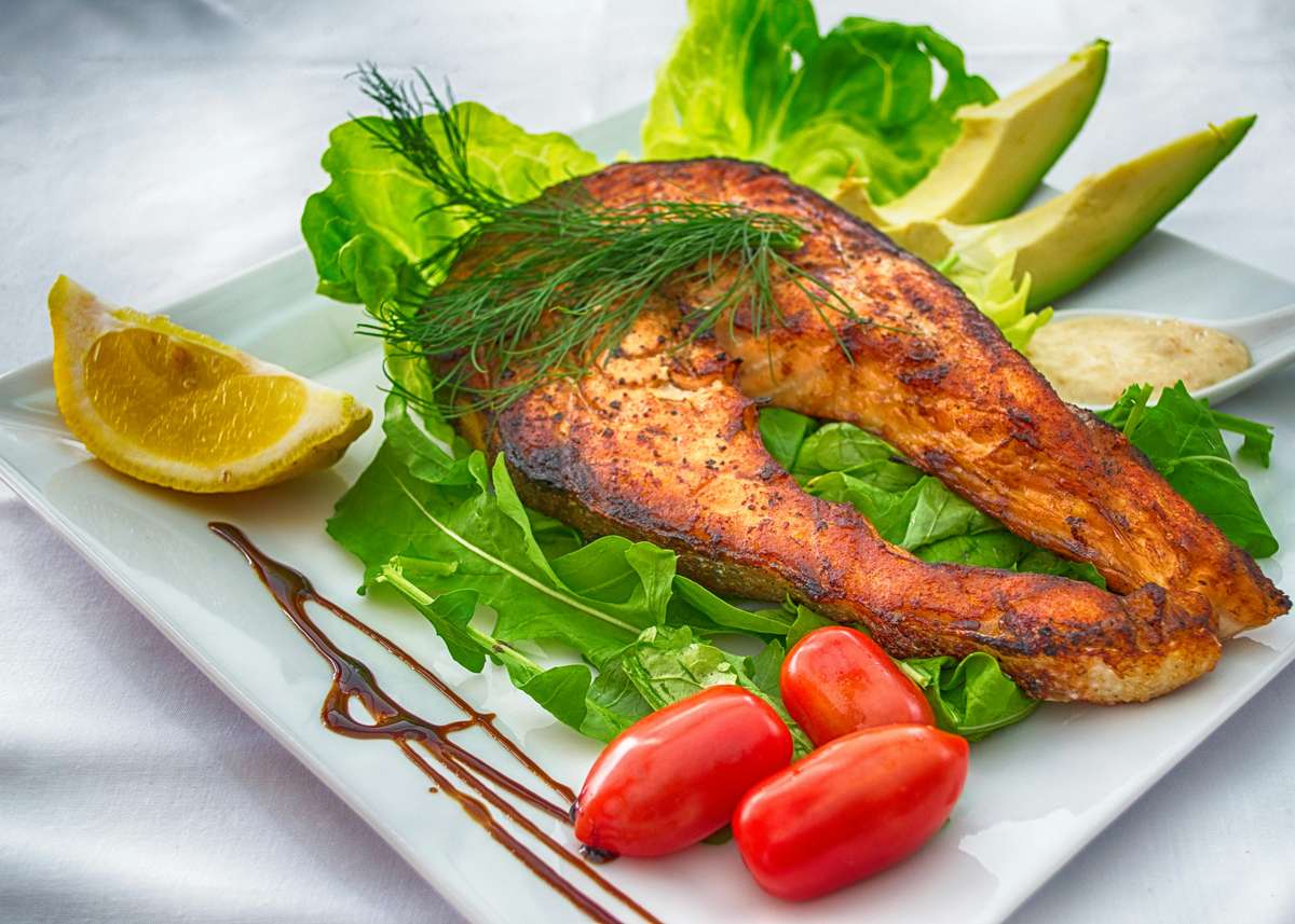 Grilled salmon steak served on a bed of fresh greens with cherry tomatoes, avocado slices, lemon wedge, and balsamic drizzle.
