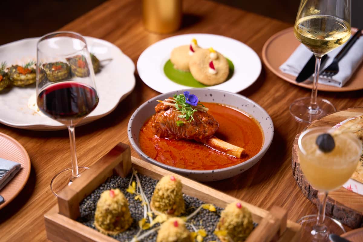 A beautifully arranged table at FITOOR featuring a rich Indian dish with tender meat in sauce, accompanied by wine, appetizers, and a vibrant cocktail, showcasing exquisite Indian cuisine.