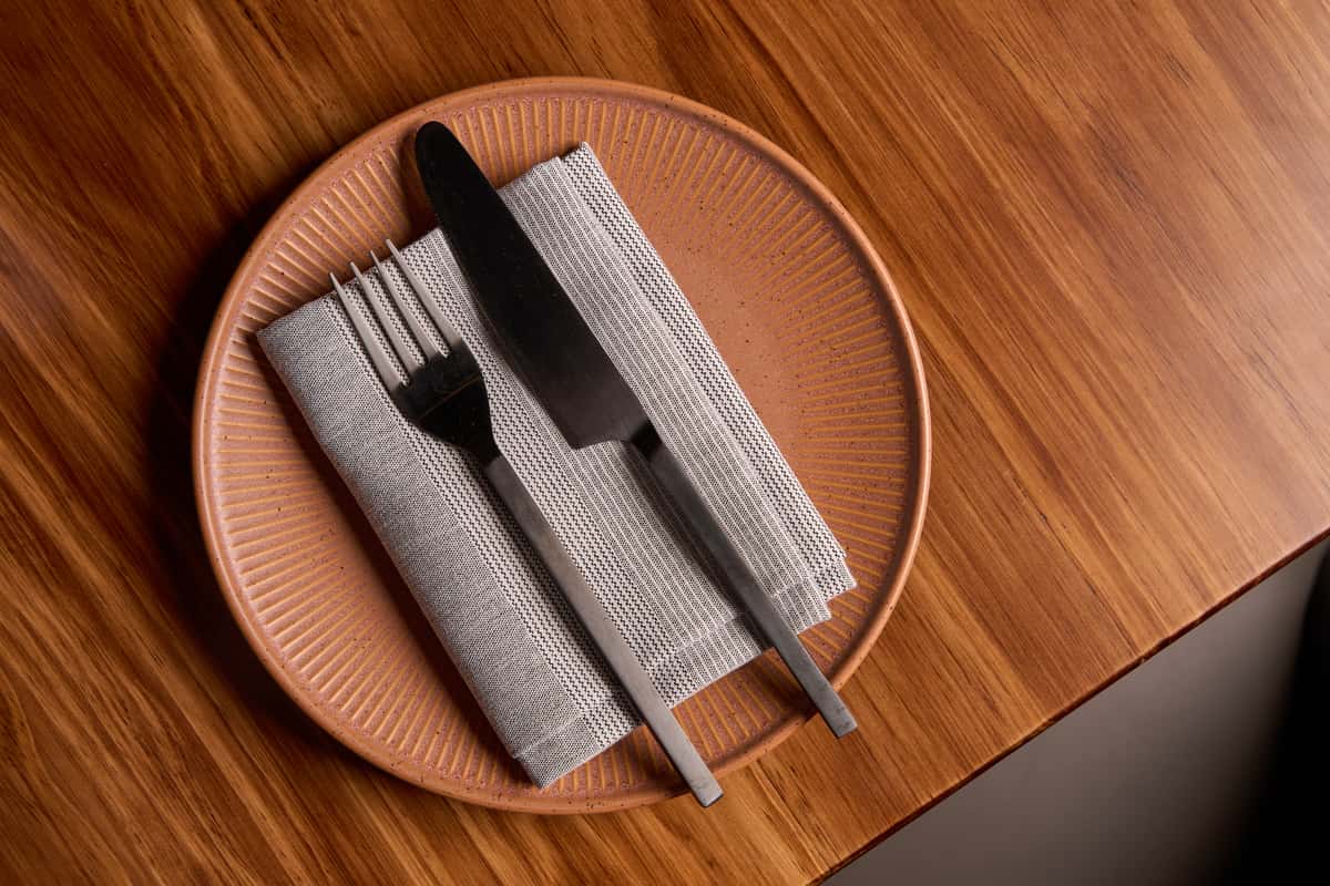 A stylish dining setup featuring a textured plate with a gray napkin, a fork, and a knife, elegantly arranged on a wooden table, perfect for a sophisticated meal experience.