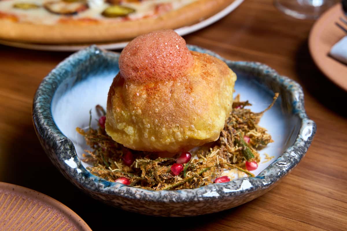 A beautifully presented dish featuring a golden, puffed pastry atop a bed of crispy herbs and pomegranate seeds, served in a decorative bowl, showcasing culinary artistry.