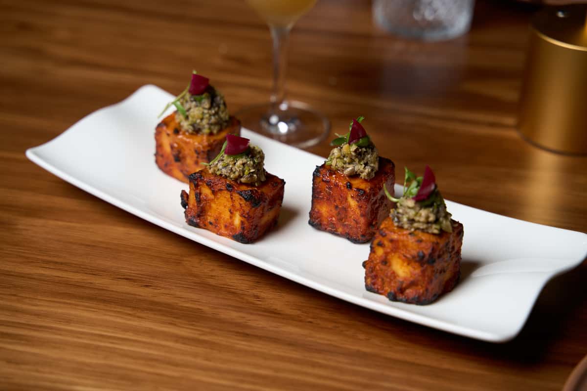 Grilled paneer cubes topped with a flavorful herb mixture and garnished with microgreens, elegantly arranged on a white plate, showcasing a gourmet Indian appetizer.