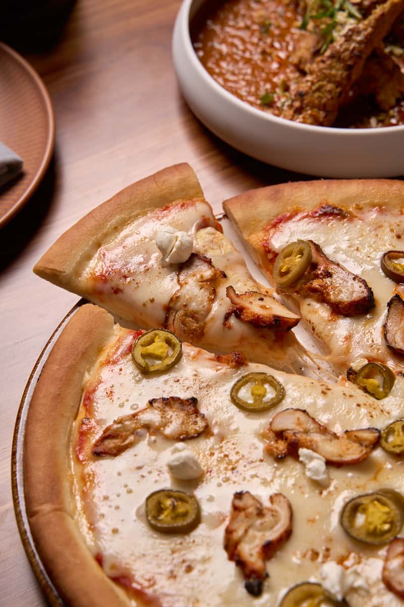 A delicious pizza featuring golden crust topped with melted cheese, grilled chicken, jalapeños, and dollops of creamy sauce, served alongside a bowl of rich sauce, perfect for sharing.