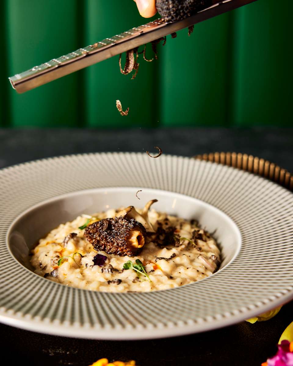 A gourmet risotto dish from FITOOR in San Jose, topped with a rich truffle being grated, garnished with herbs and edible flowers, showcasing exquisite culinary craftsmanship.