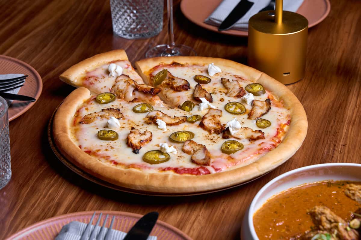A fusion pizza topped with spiced chicken, jalapeños, and dollops of cream, served alongside a bowl of rich Indian curry, showcasing a blend of flavors and culinary creativity.