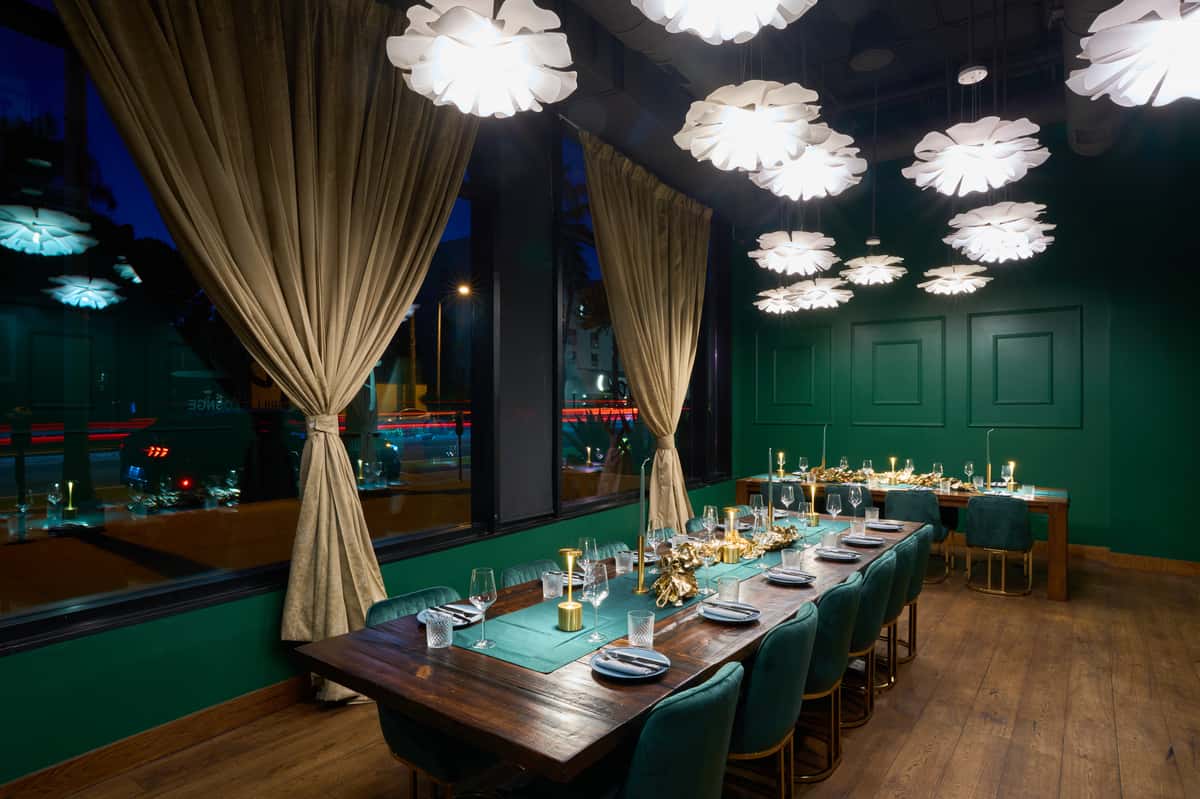 An elegant dining room at FITOOR, featuring a long wooden table set for reservations, adorned with stylish tableware, soft lighting, and rich green walls, creating a cozy atmosphere.