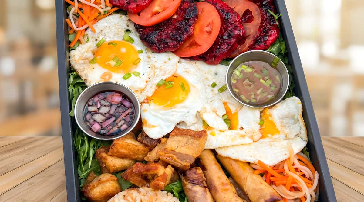 A platter with sunny-side-up eggs, red meat slices with tomatoes, fried pork belly, egg rolls, rice, and sauces.