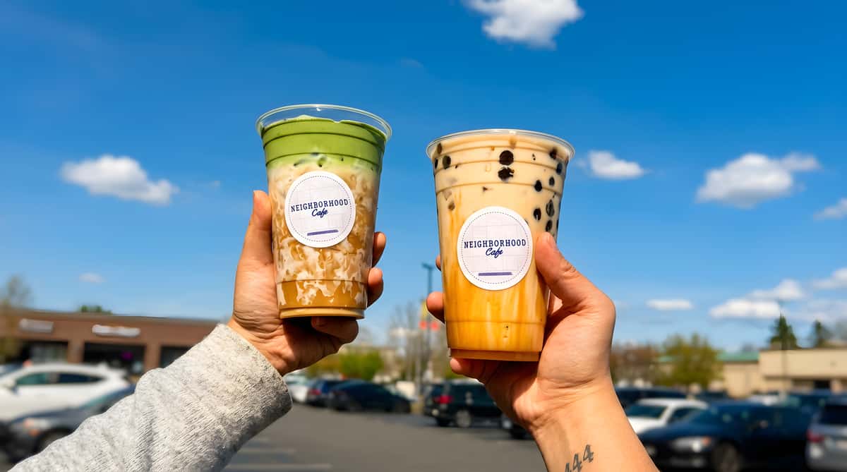 Two hands hold up plastic cups of iced beverages: a green-topped coffee and a boba tea, against a blue sky.