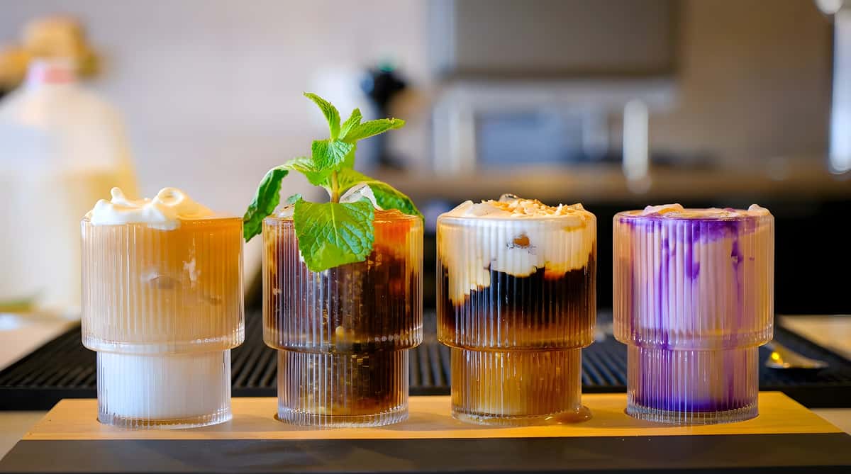 Four layered drinks in ribbed glasses: one with whipped cream, one with mint, one with white foam and coconut, and one purple and white.