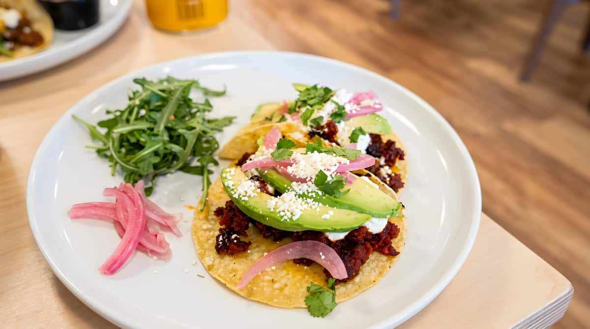 Tacos with avocado slices, red onions, crumbled cheese, and cilantro, served with a side of arugula.