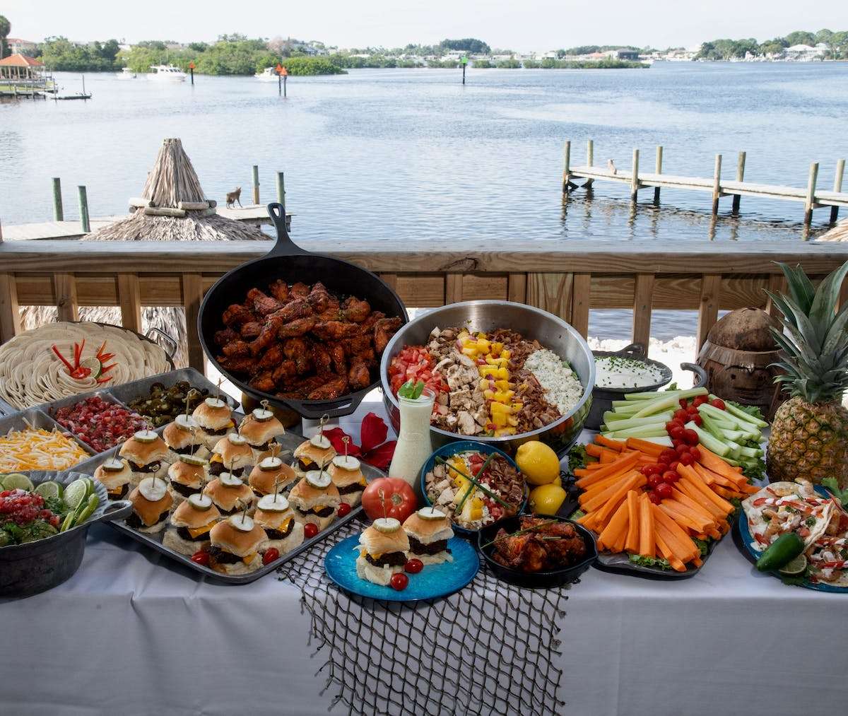 Waterfront catering buffet featuring sliders, wings, fresh vegetables, taco toppings, and party appetizers with bay views