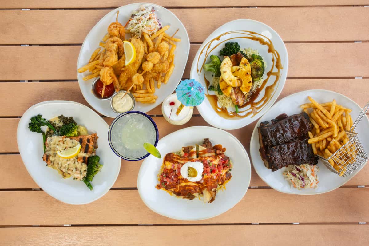 Overhead view of seafood platters, barbecue ribs, tropical cocktails, and waterfront restaurant favorites served family-style on a wooden table