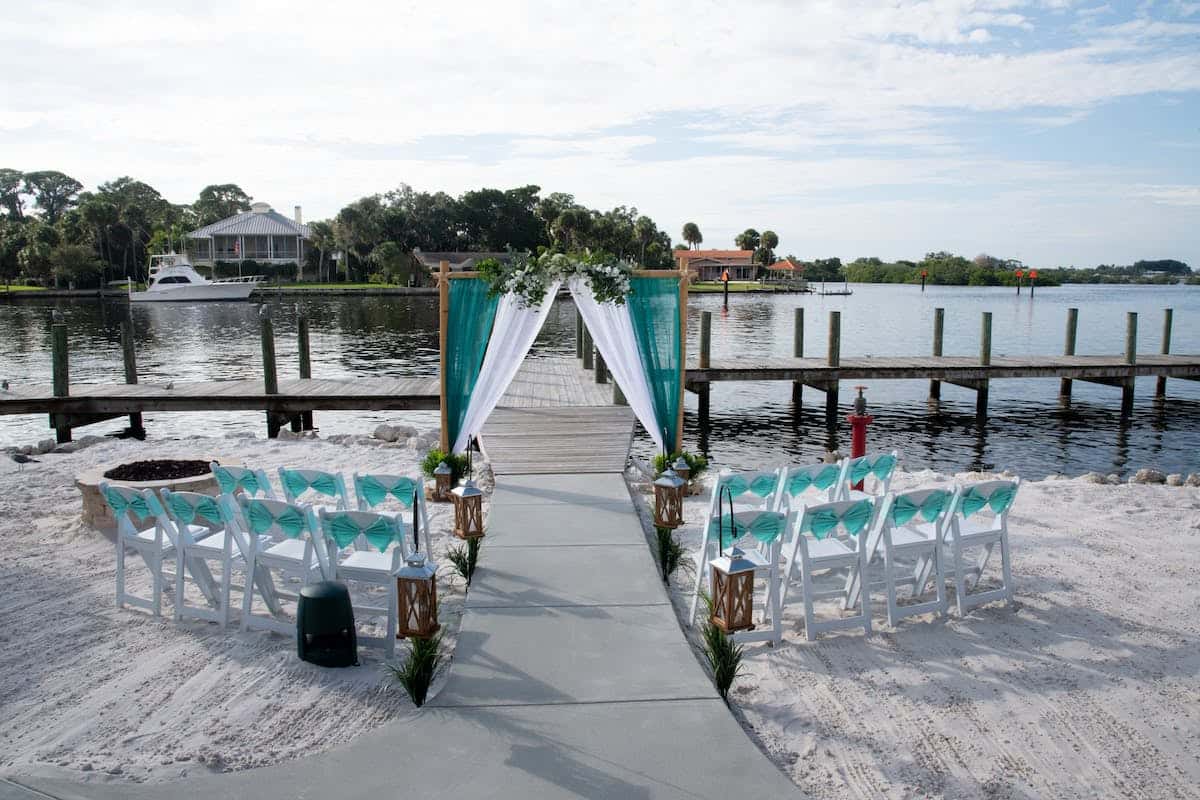 Waterfront beach wedding ceremony setup with white chairs, turquoise draping, and dockside views along the bay