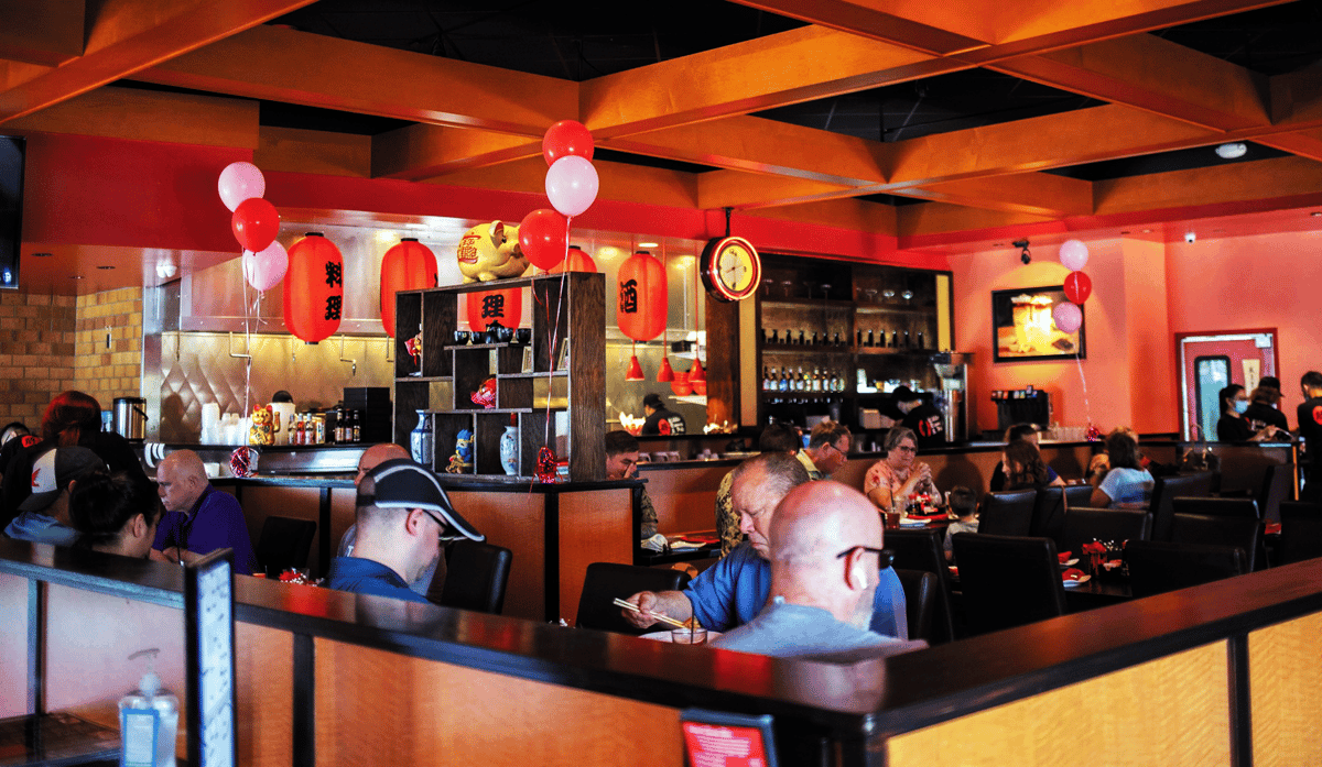 Busy Asian restaurant dining room with red lanterns, warm decor, open kitchen, and guests seated at tables