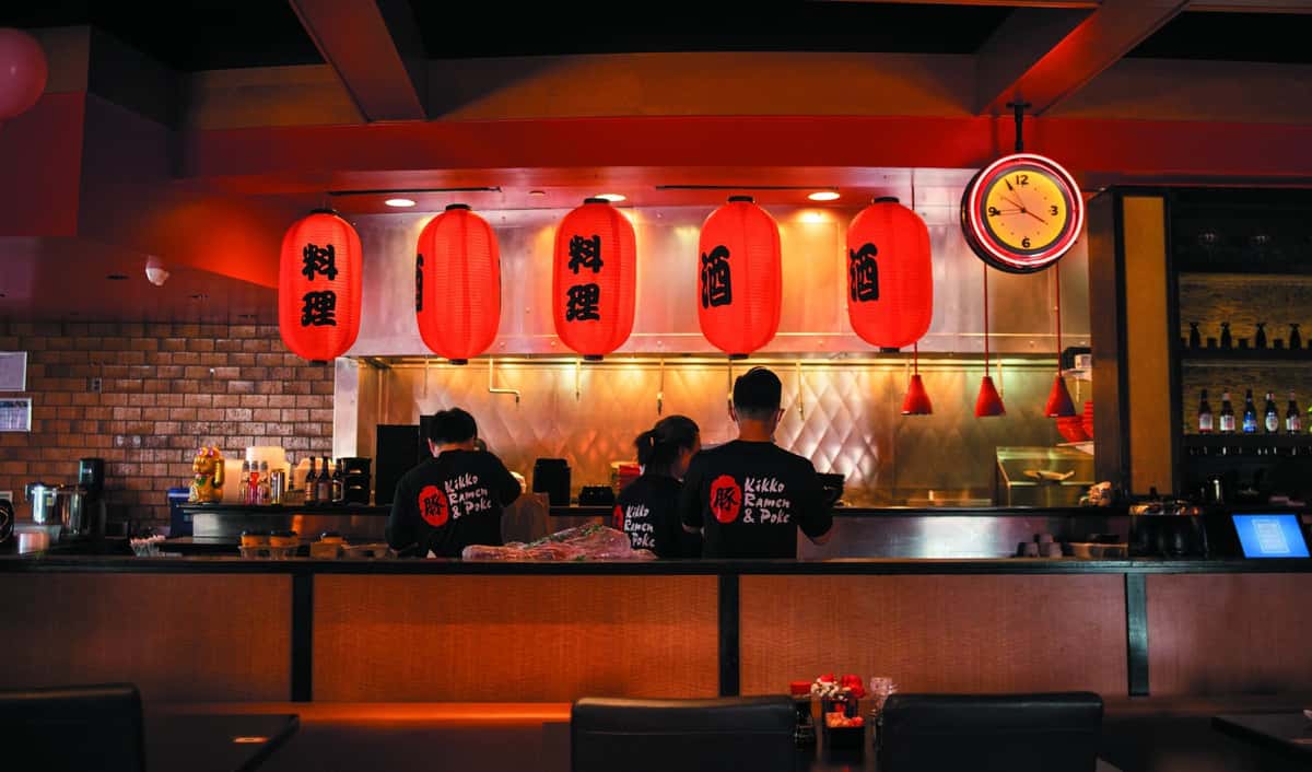 Japanese ramen restaurant interior with red lanterns, open kitchen, and chefs preparing food behind the counter