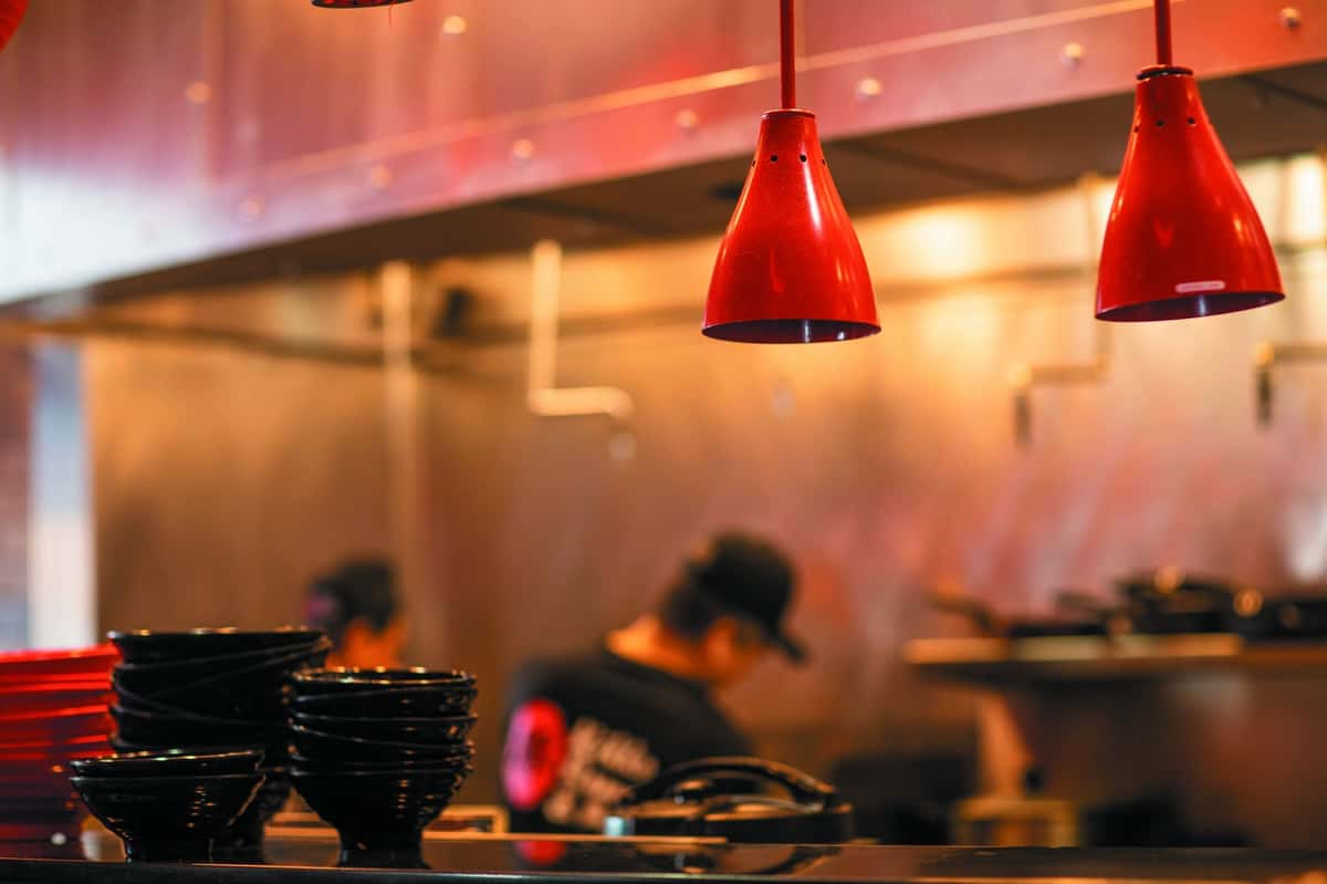Open kitchen at an Asian restaurant with red pendant lights, stacked bowls, and chefs preparing food