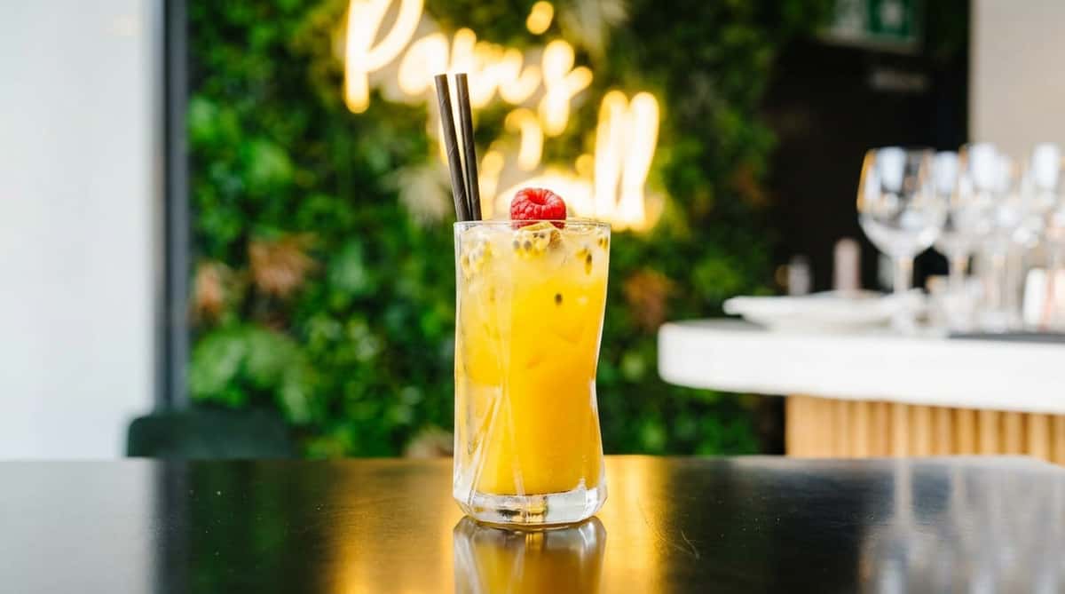 Yellow cocktail with ice, passion fruit seeds, a black straw, and a red fruit garnish, on a dark table.