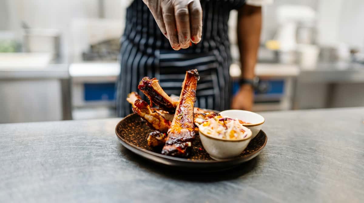 Glazed pork ribs on a plate with coleslaw and sauce, being garnished by a gloved hand.