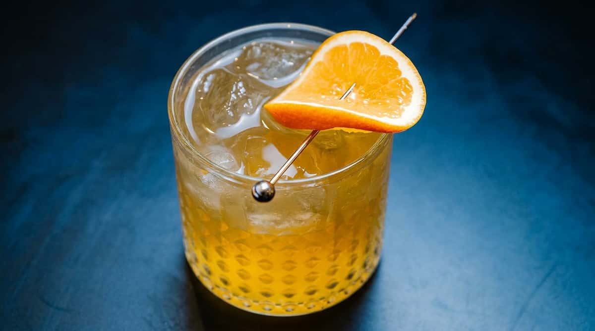 Old Fashioned cocktail with ice and an orange slice garnish on a dark surface.