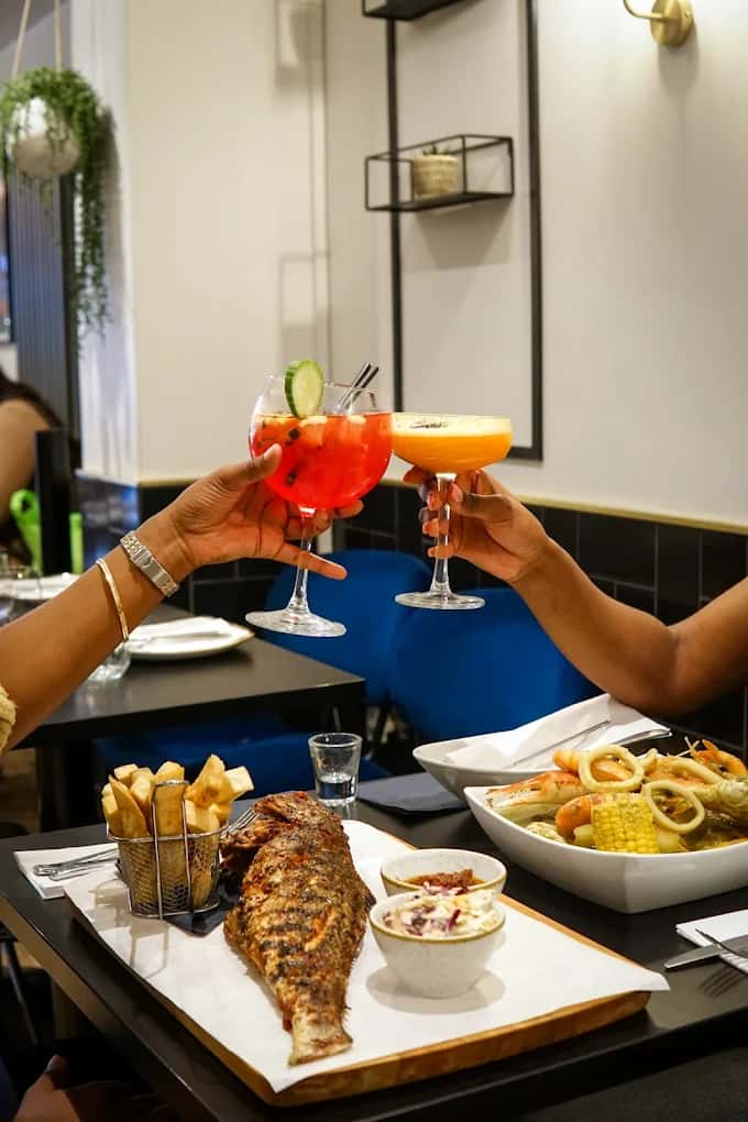 Friends toasting colorful cocktails over a restaurant table with grilled whole fish, fries, and seafood dishes in a modern dining setting
