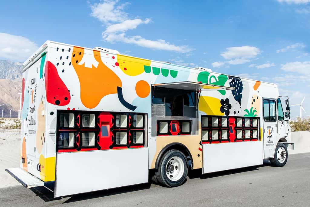 Whimsically decorated food truck with open serving windows, patterned with abstract cow spots and flowers, parked under a clear sky with mountains in the background.