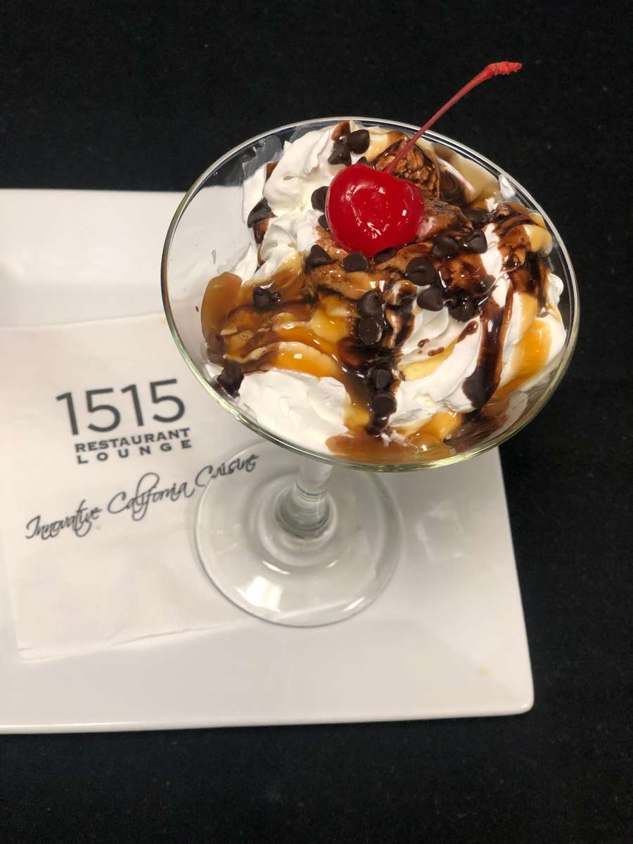 A decadent dessert is served in a glass dish, featuring scoops of creamy ice cream topped with rich chocolate and caramel drizzle, crunchy nuts, and a bright red cherry on top. The indulgent presentation makes it a perfect sweet finish to a meal.