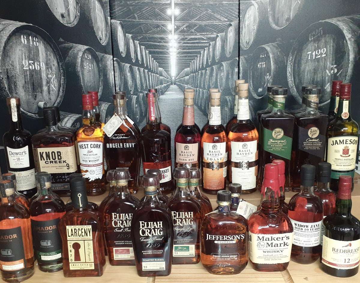 Extensive collection of premium whiskey and bourbon bottles, including Elijah Craig, Basil Hayden, Knob Creek, and Jameson, displayed in front of a black-and-white barrel warehouse mural, ideal for a whiskey bar or tasting room setting.