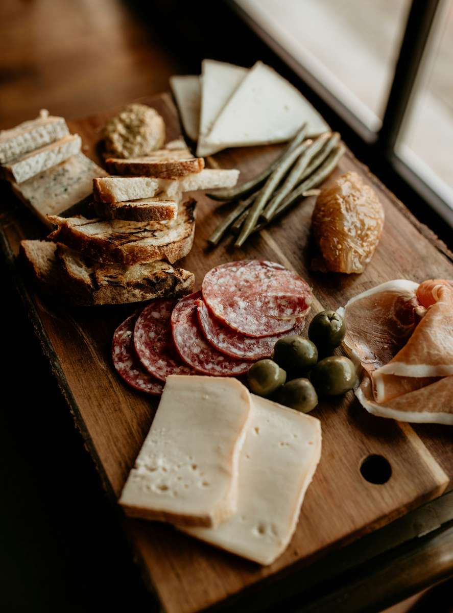 Rustic charcuterie board with sliced salami, prosciutto, cheeses, grilled bread, olives, pickled vegetables, and whole grain mustard, set beside natural window light for a cozy dining ambiance.
