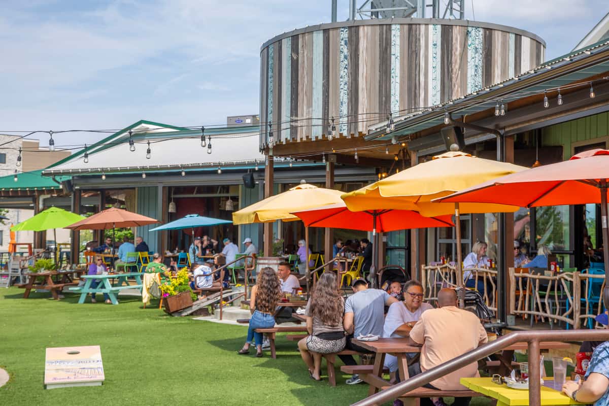 Crowded patio at Crooked Hammock Brewery with colorful umbrellas, cornhole game, and diners enjoying food and drinks.