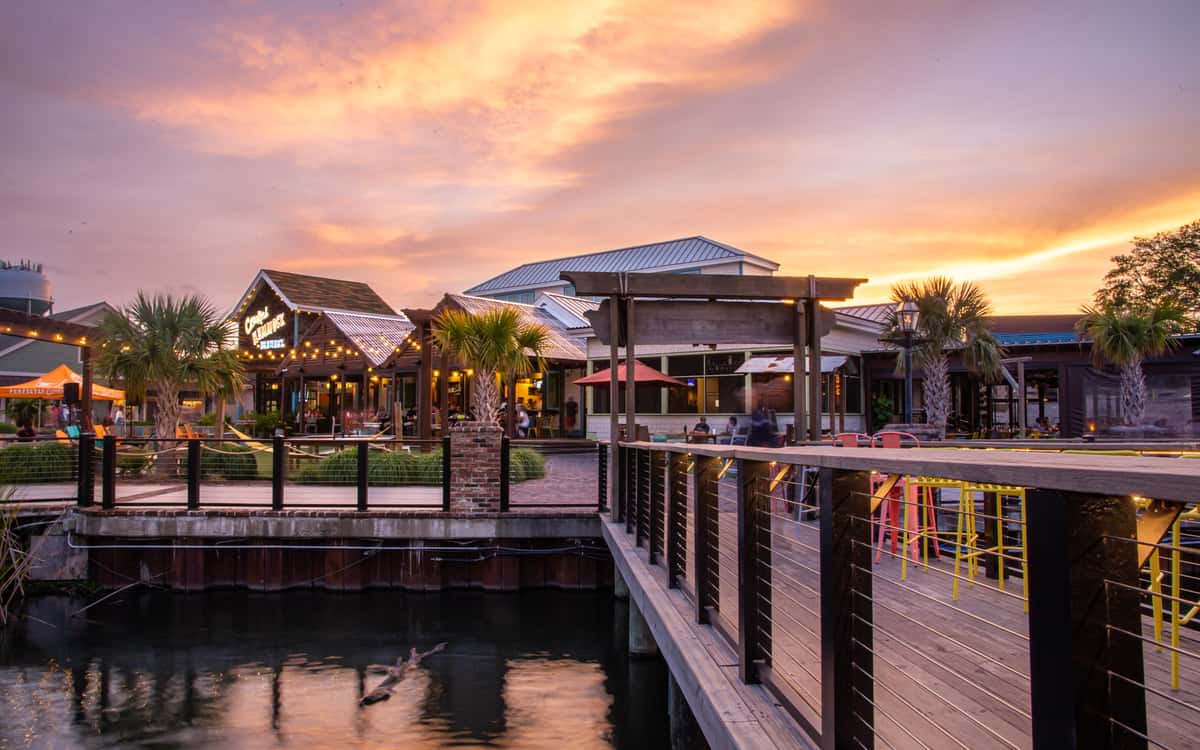 Waterfront view of Crooked Hammock Brewery at sunset with glowing string lights, palm trees, and boardwalk reflections.