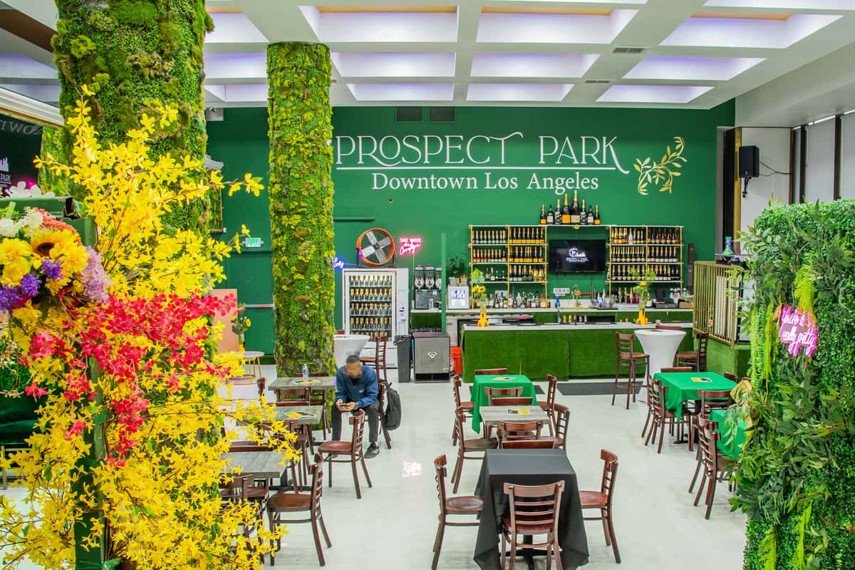 This vibrant and lush venue, Prospect Park Downtown Los Angeles, features a nature-inspired aesthetic with green faux grass, foliage-covered columns, and colorful floral arrangements.