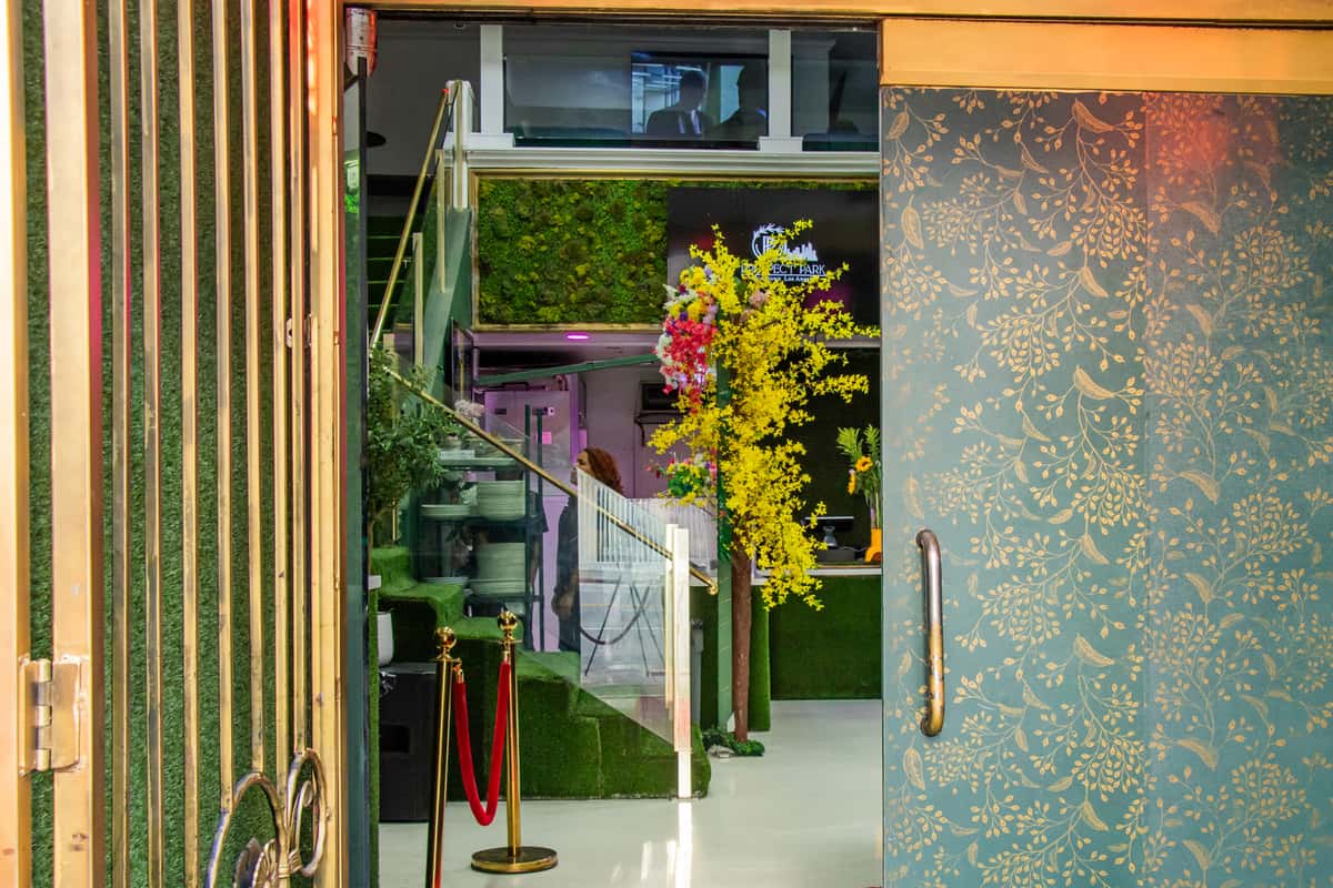 Open doorway revealing a vibrant, greenery-filled interior with a staircase, floral decor, and Prospect Park signage in the background.
