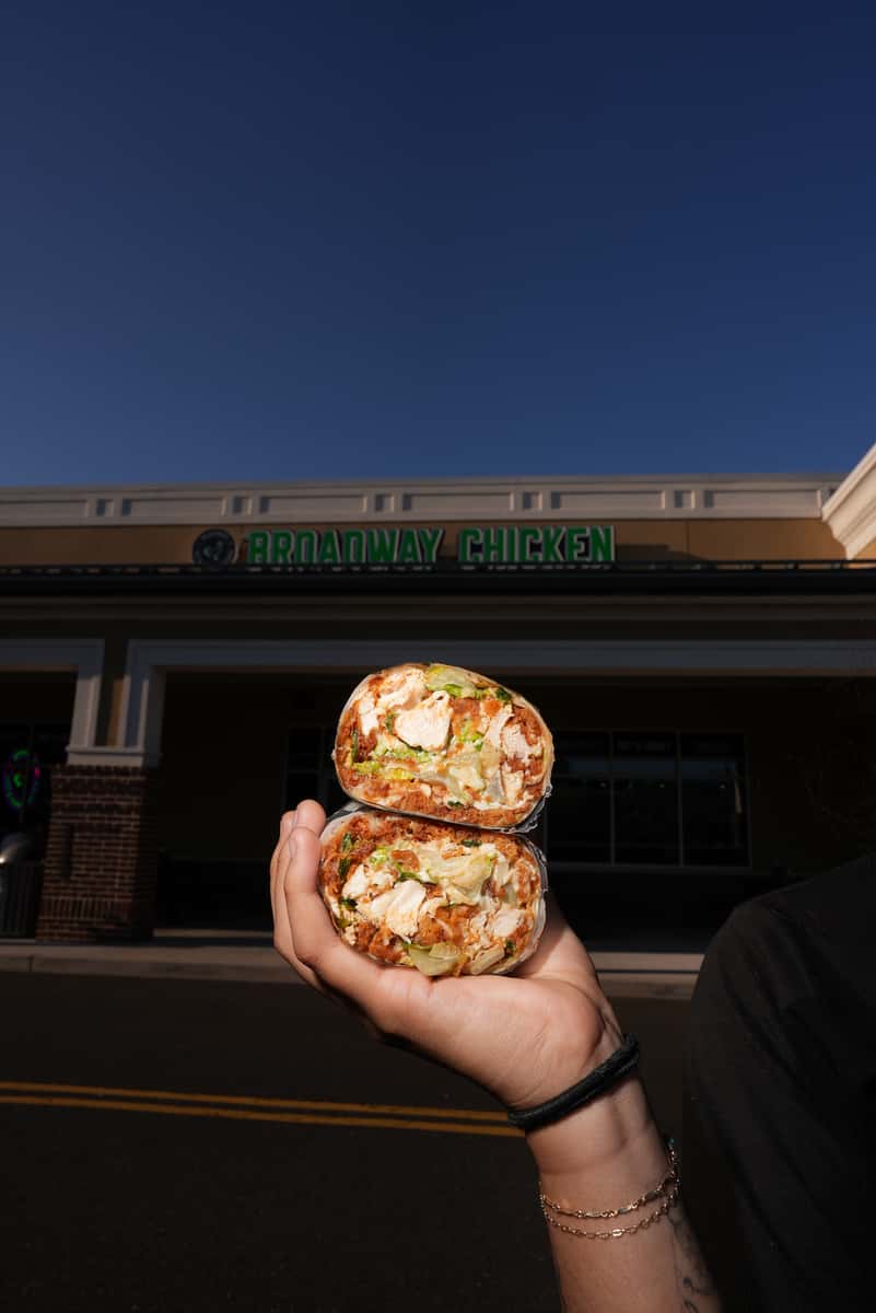 Hand holding a sandwich wrap in front of a Broadway Chicken location