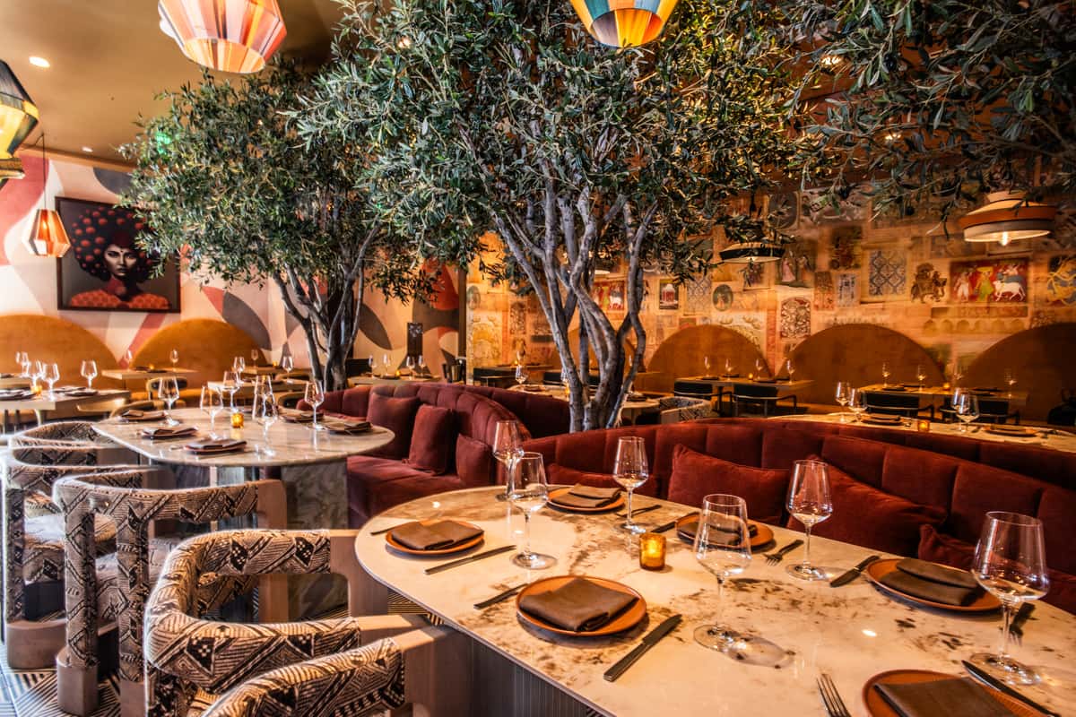 Upscale restaurant interior with indoor olive trees, marble tables, velvet banquette seating, and warm ambient lighting