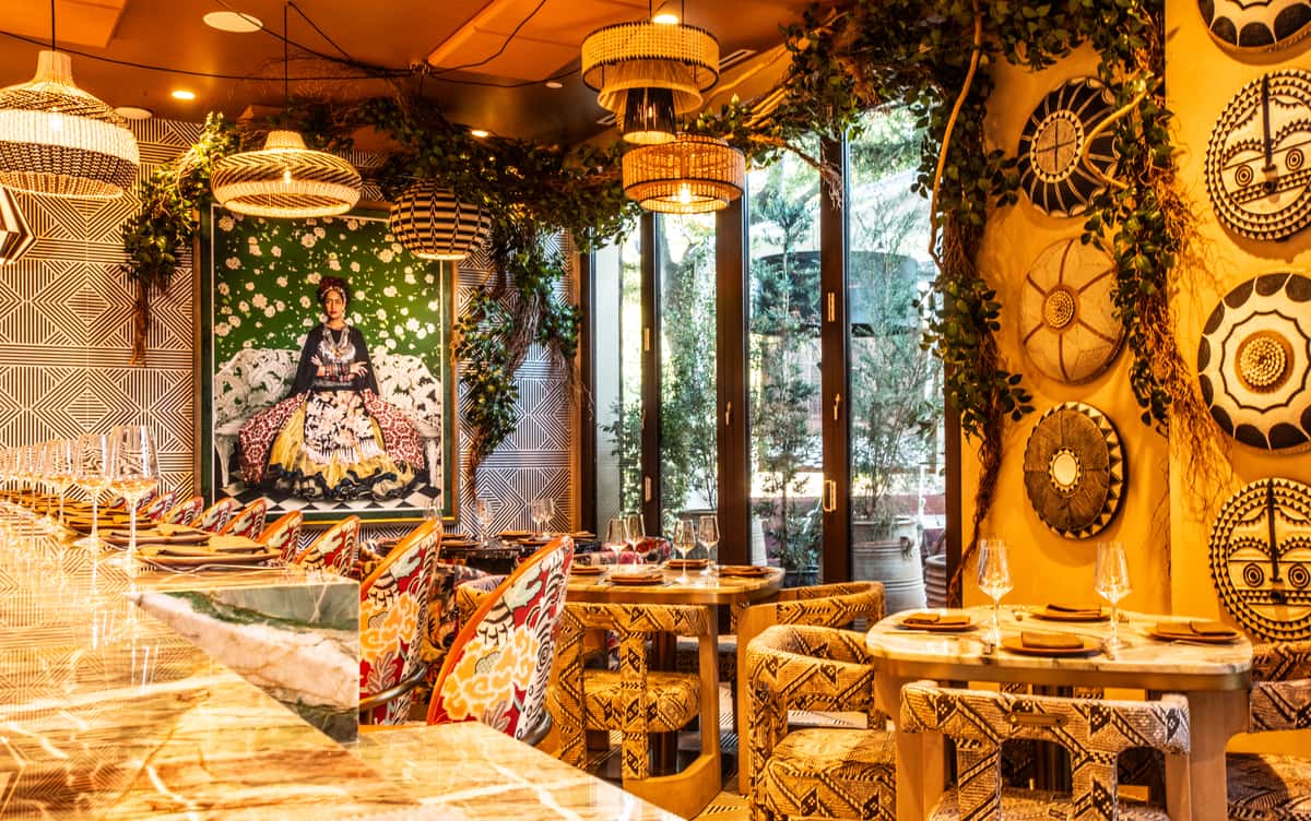 Warm, eclectic restaurant dining room with patterned chairs, woven pendant lights, lush greenery, and vibrant global-inspired decor