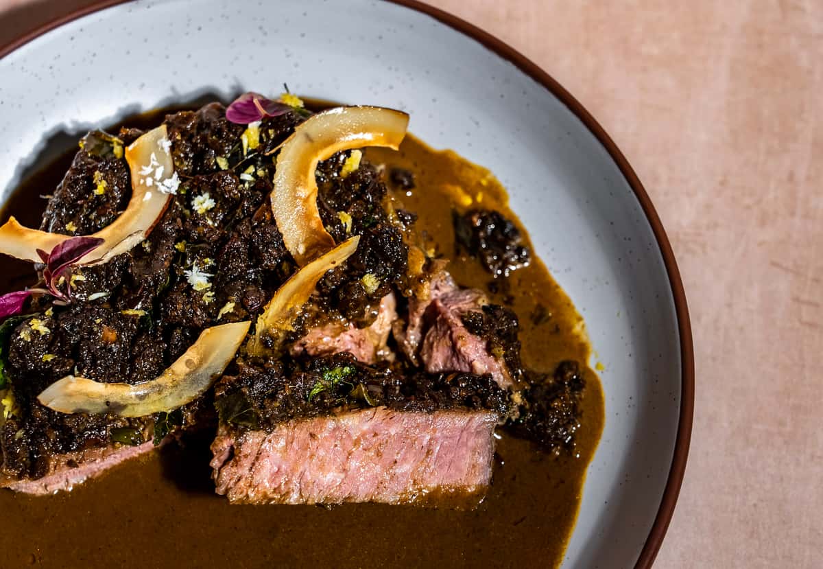 Medium-rare sliced steak in rich brown sauce with caramelized onions, herbs, and citrus garnish in a modern fine dining bowl